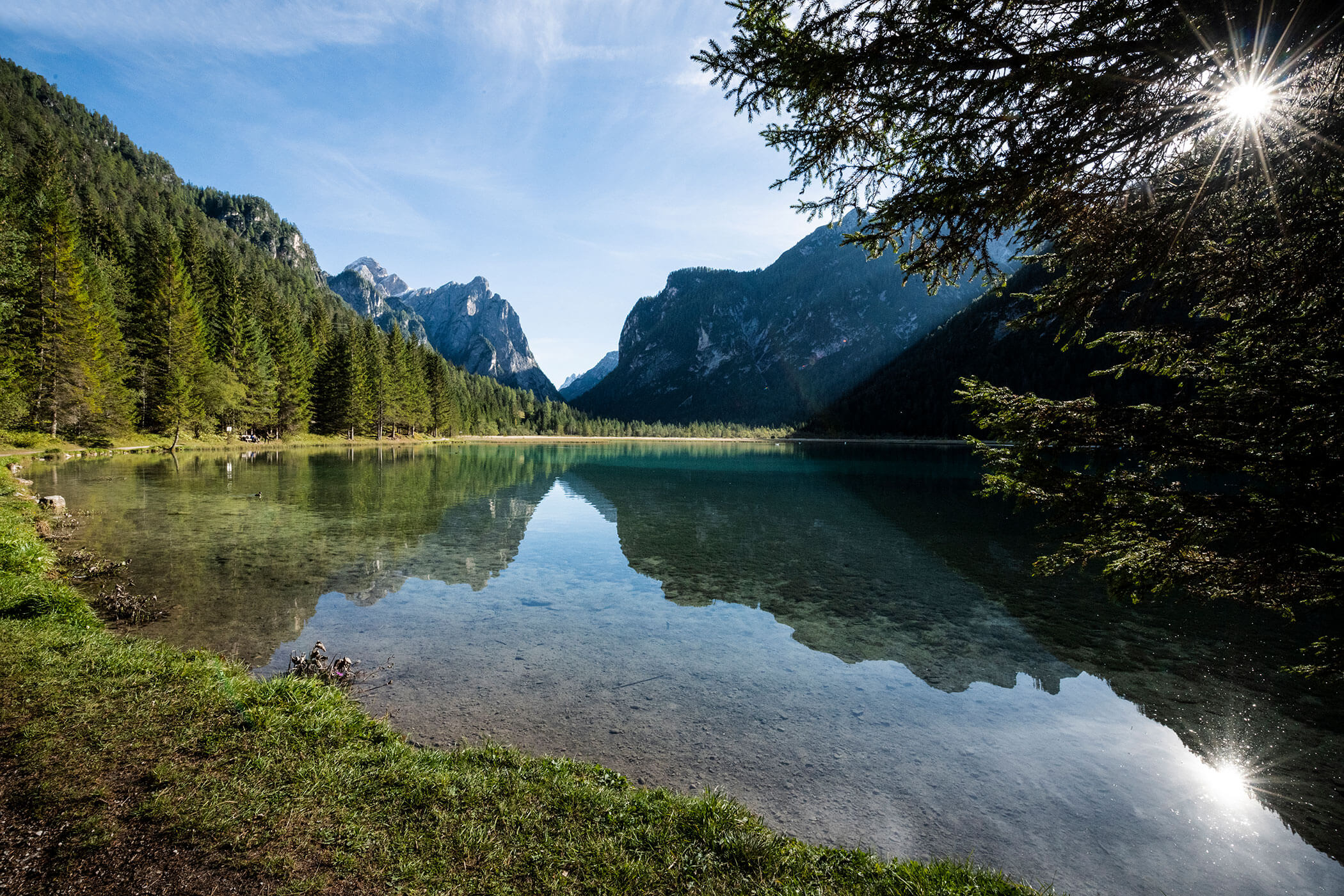Clear mountain lake with trees and distant peaks, sun shines through branches, sky and mountains reflected in water. - Hotel Innerhofer