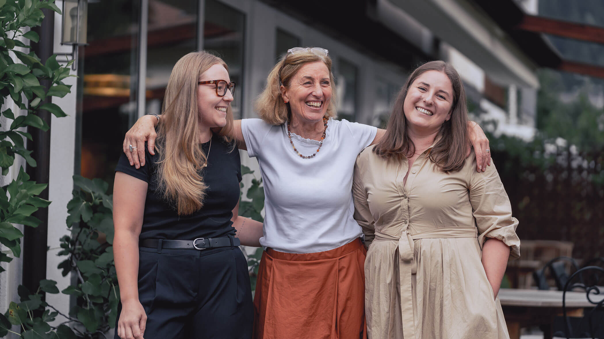 Three women stand outside, smiling and laughing together, with the woman in the middle hugging the others. - Hotel Innerhofer