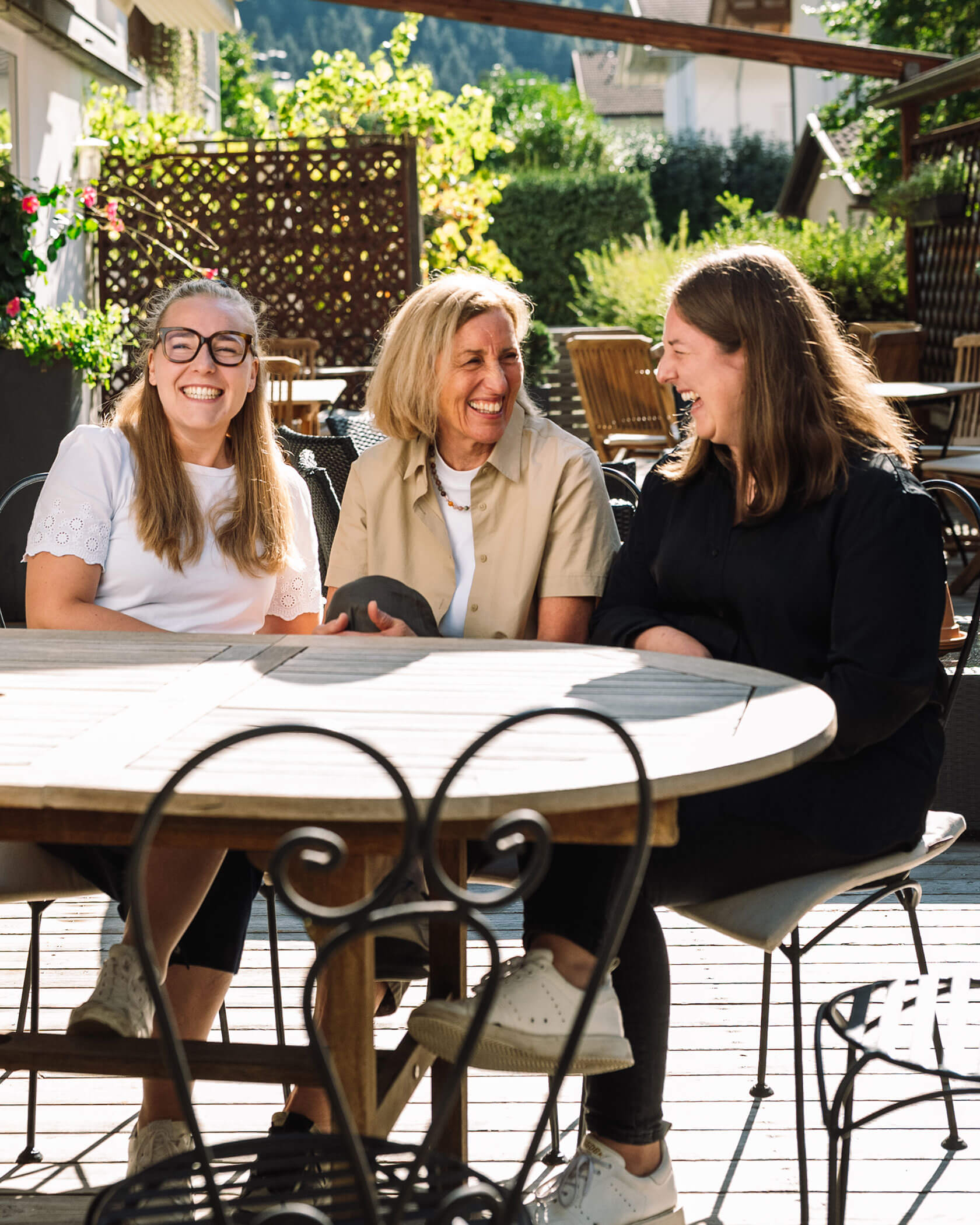 Three women sit at an outdoor table, smiling and laughing together on a sunny day. - Hotel Innerhofer