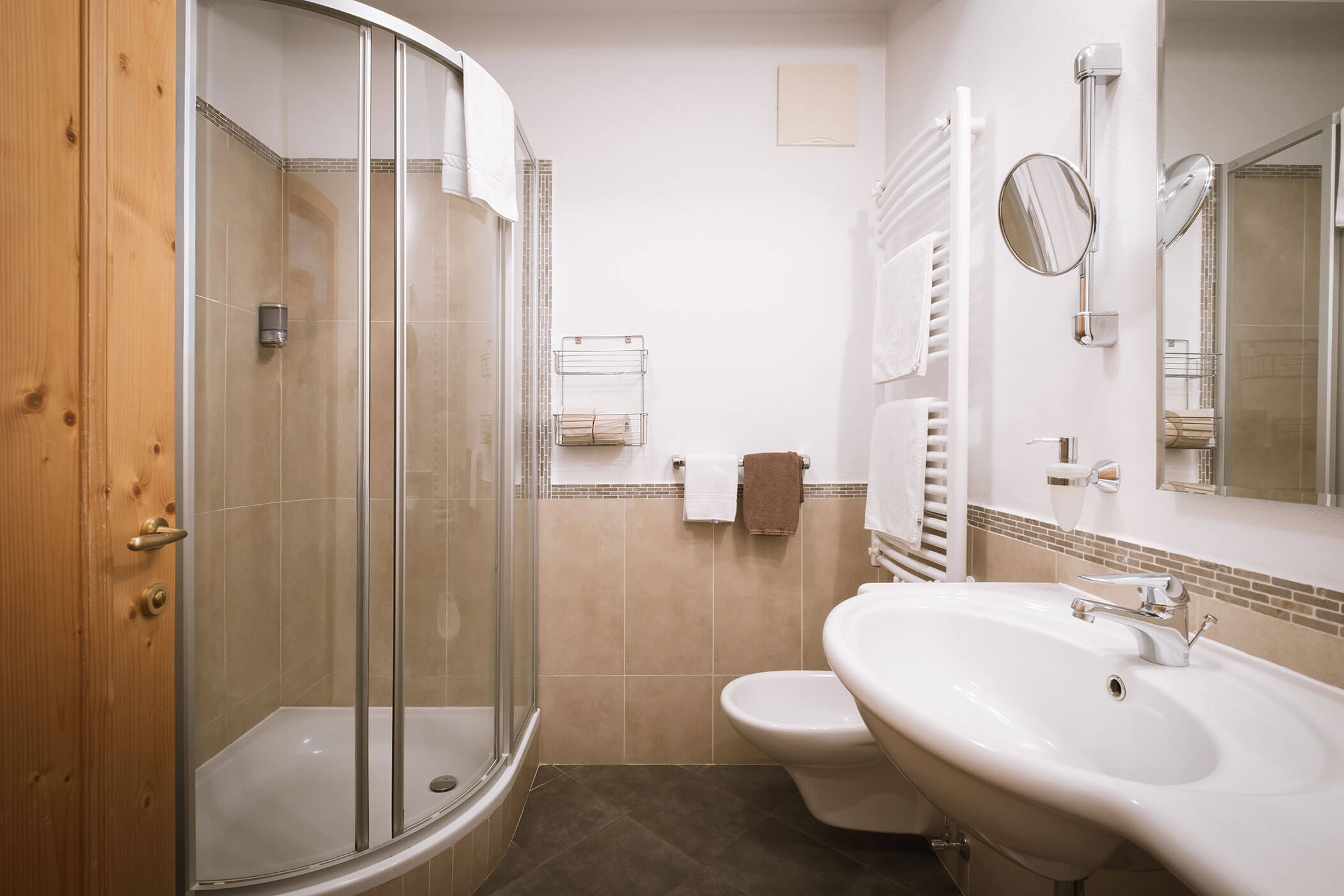 Bagno moderno con doccia in vetro, lavandino bianco, WC, portasciugamani e pareti piastrellate beige. - Hotel Innerhofer