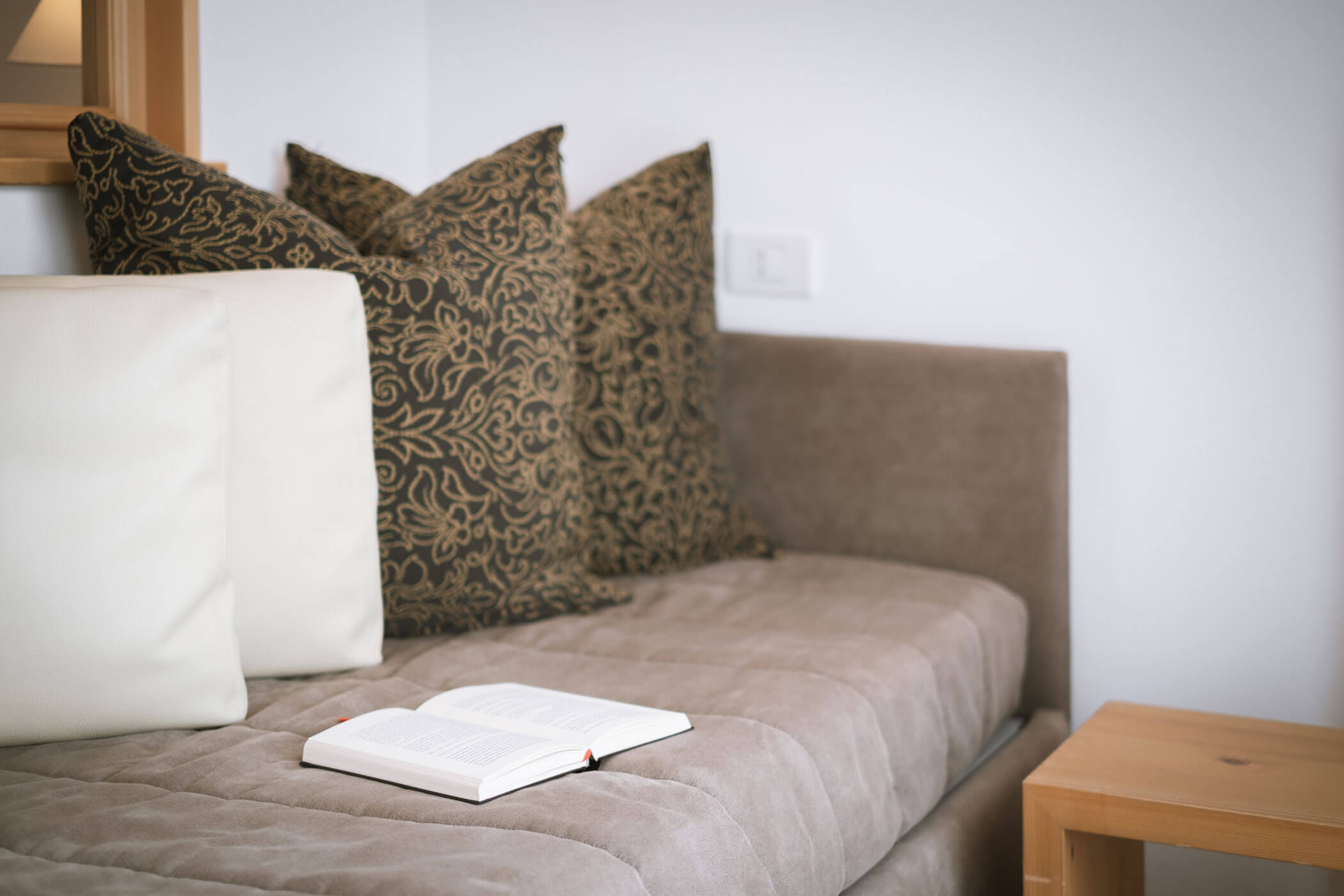 Un divano beige con cuscini a fantasia e un libro aperto appoggiato sulla seduta accanto a un tavolo di legno. - Hotel Innerhofer