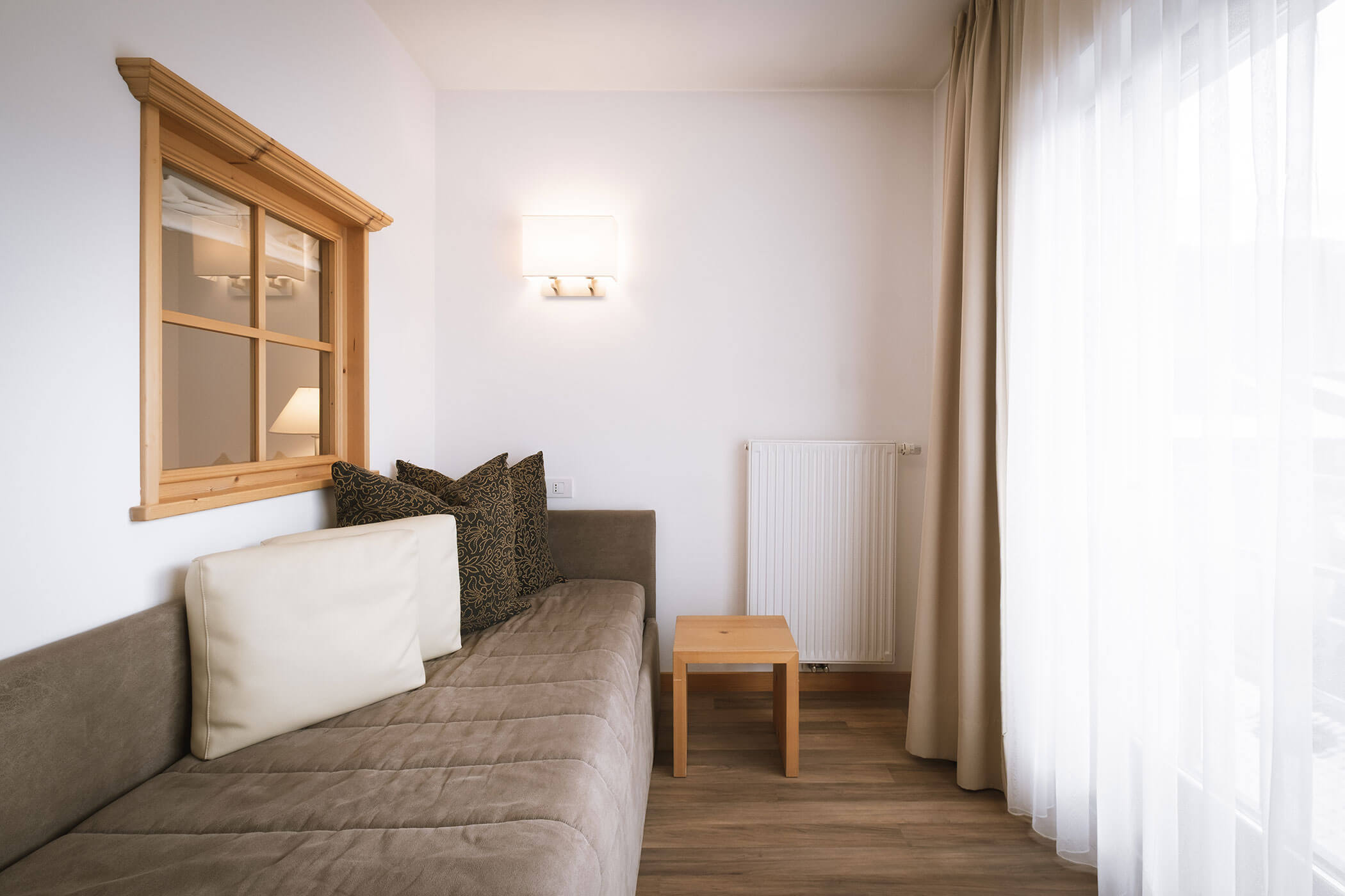 Soggiorno minimalista con divano marrone, cuscini, tavolino in legno e tende trasparenti vicino a una grande finestra. - Hotel Innerhofer