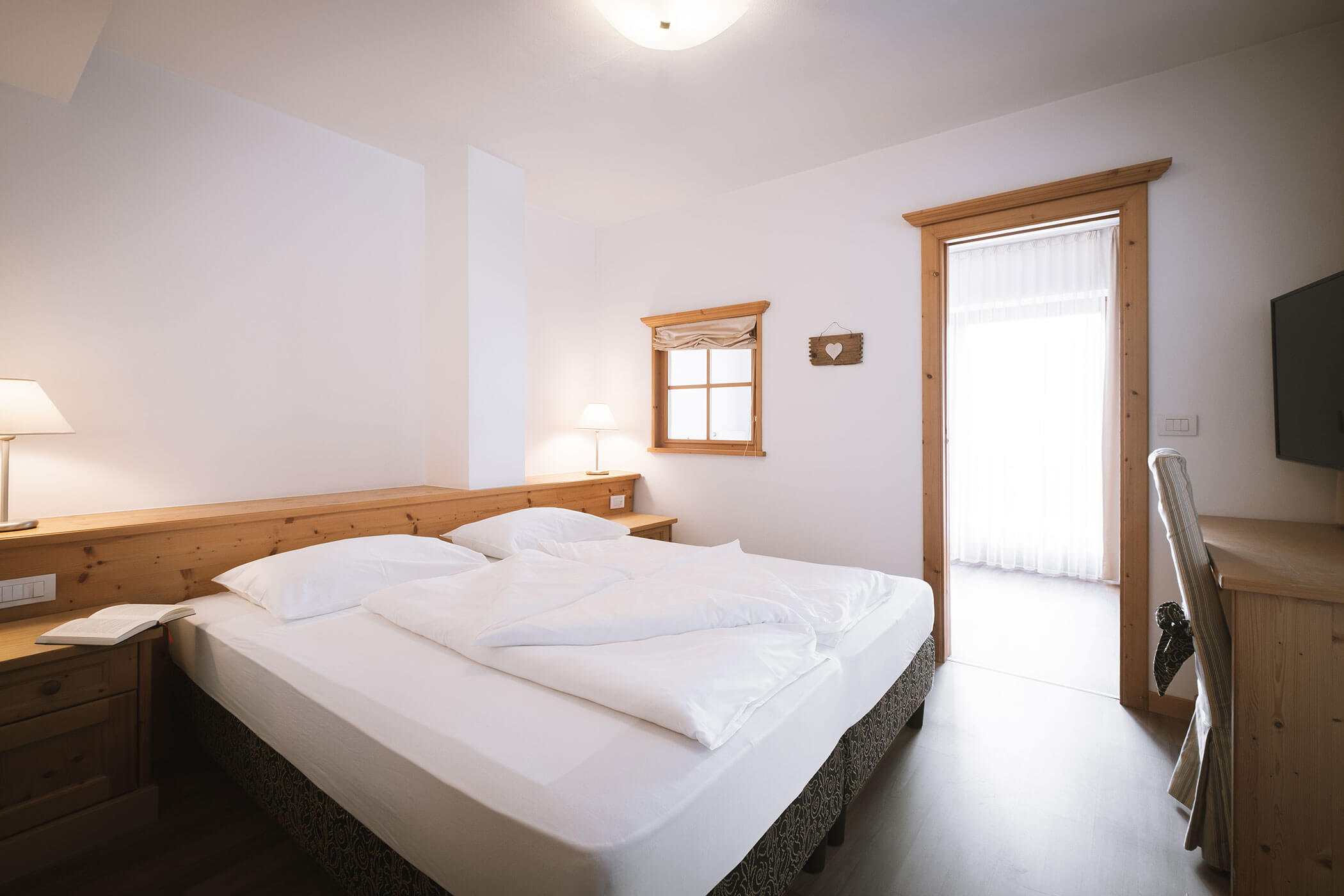 Camera d'albergo luminosa e accogliente con letto matrimoniale, mobili in legno, lampade e una porta aperta su un balcone illuminato dal sole. - Hotel Innerhofer