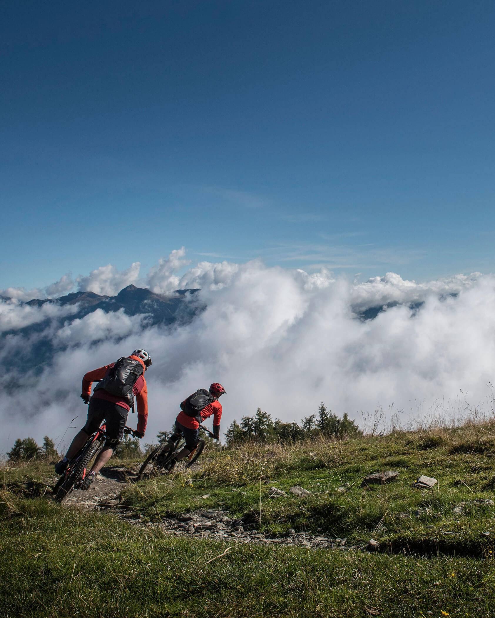 Zwei Radfahrer fahren mit ihren Mountainbikes auf einem grasbewachsenen Weg mit Wolken und Bergen im Hintergrund. - Hotel Innerhofer
