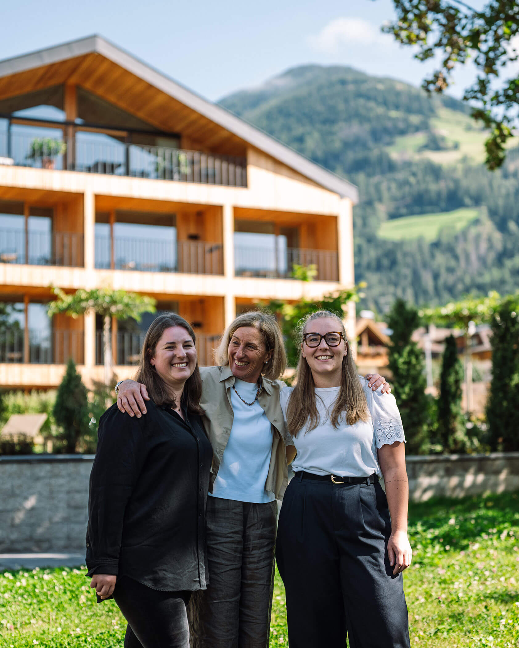Drei Frauen lächeln und posieren zusammen vor einem Holzgebäude mit Bergen im Hintergrund. - Hotel Innerhofer