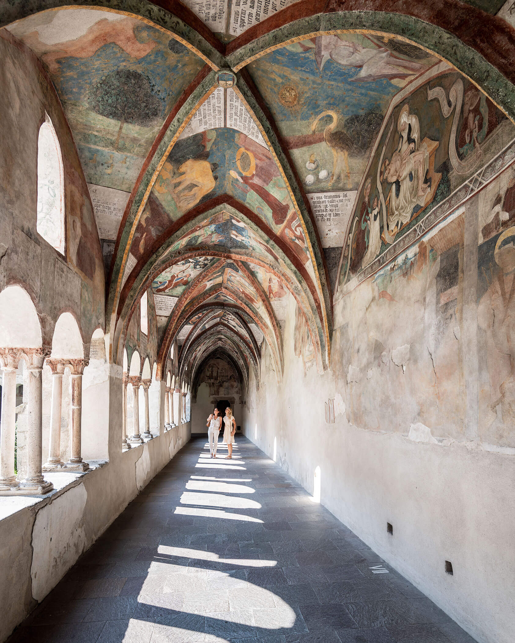 People walk down a sunlit, frescoed cloister with arched ceilings and stone floor. - Hotel Innerhofer
