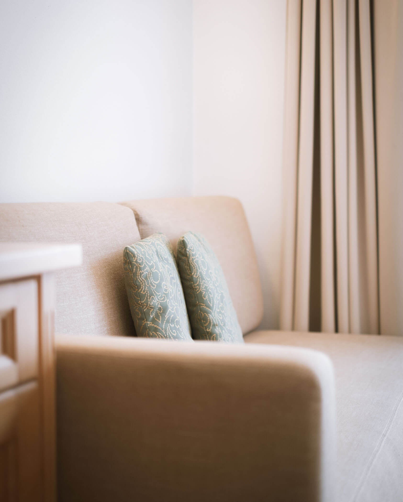 Hellbraunes Sofa mit zwei gemusterten grünen Kissen neben beigen Vorhängen in einem sanft beleuchteten Raum. - Hotel Innerhofer