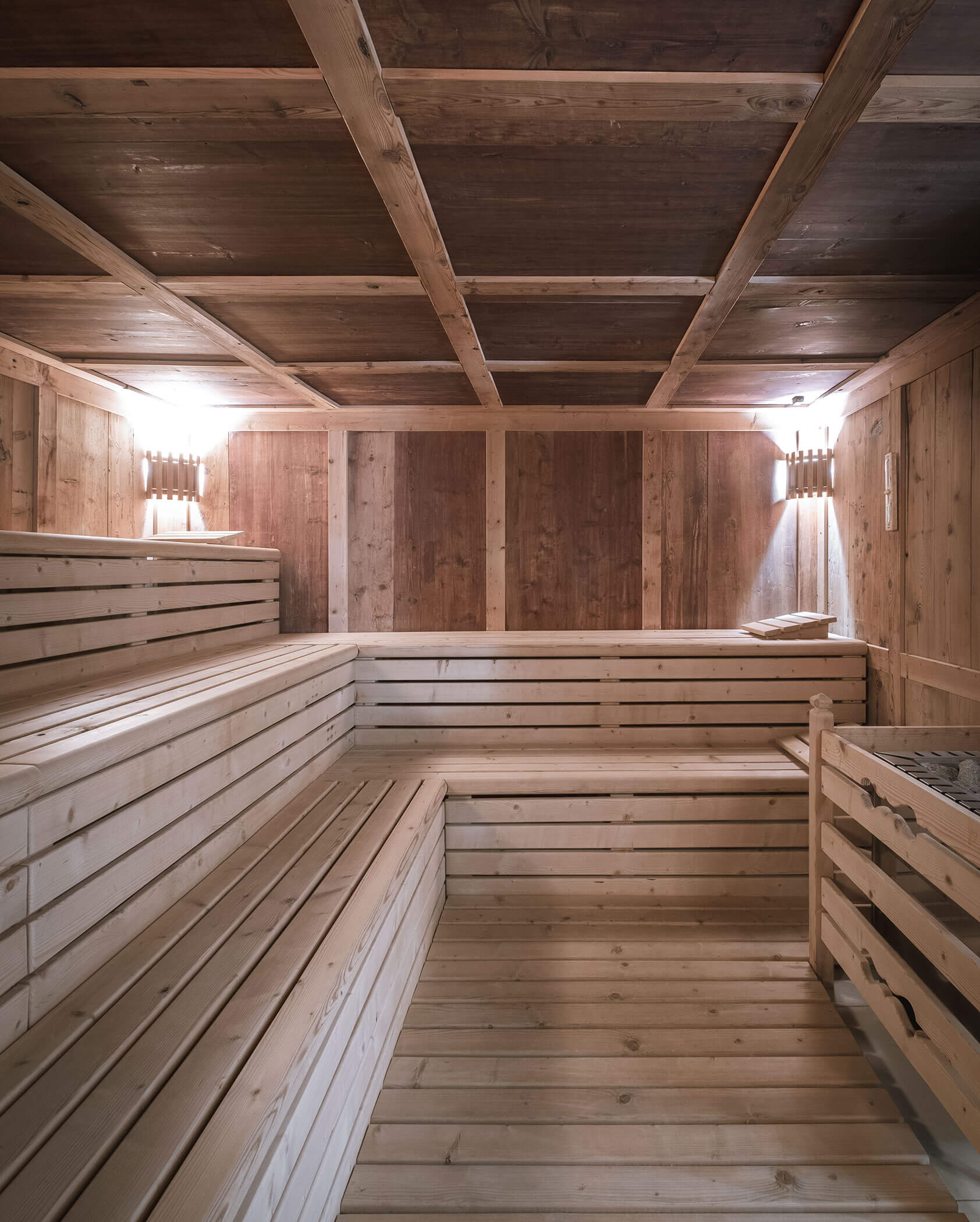 Holzverkleidete Sauna mit Stufenbänken, warmer Beleuchtung und einem Ofen auf der rechten Seite. - Hotel Innerhofer