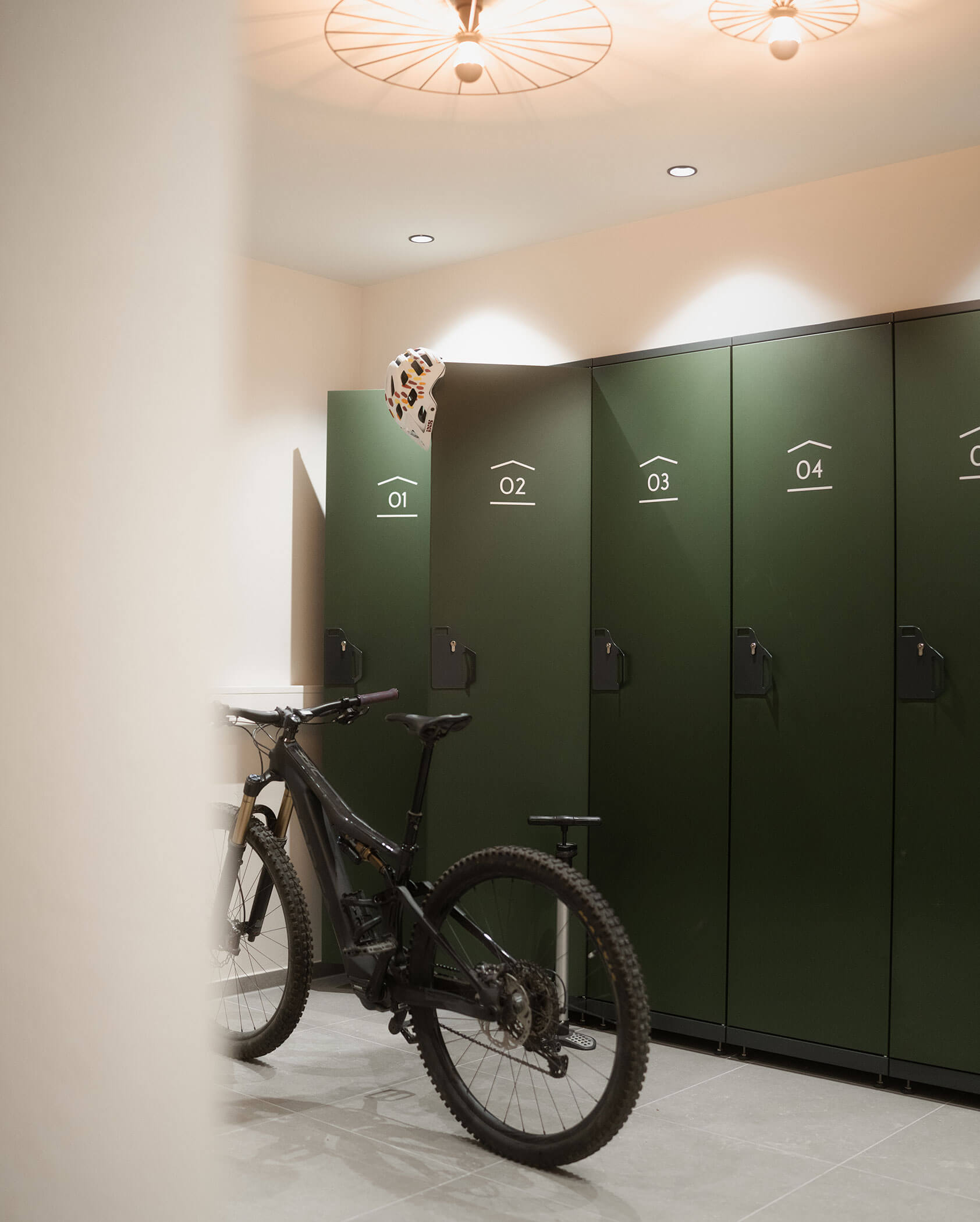 Una bicicletta nera è parcheggiata davanti agli armadietti verdi numerati da 01 a 04; sull'armadietto 02 è appoggiato un casco. - Hotel Innerhofer