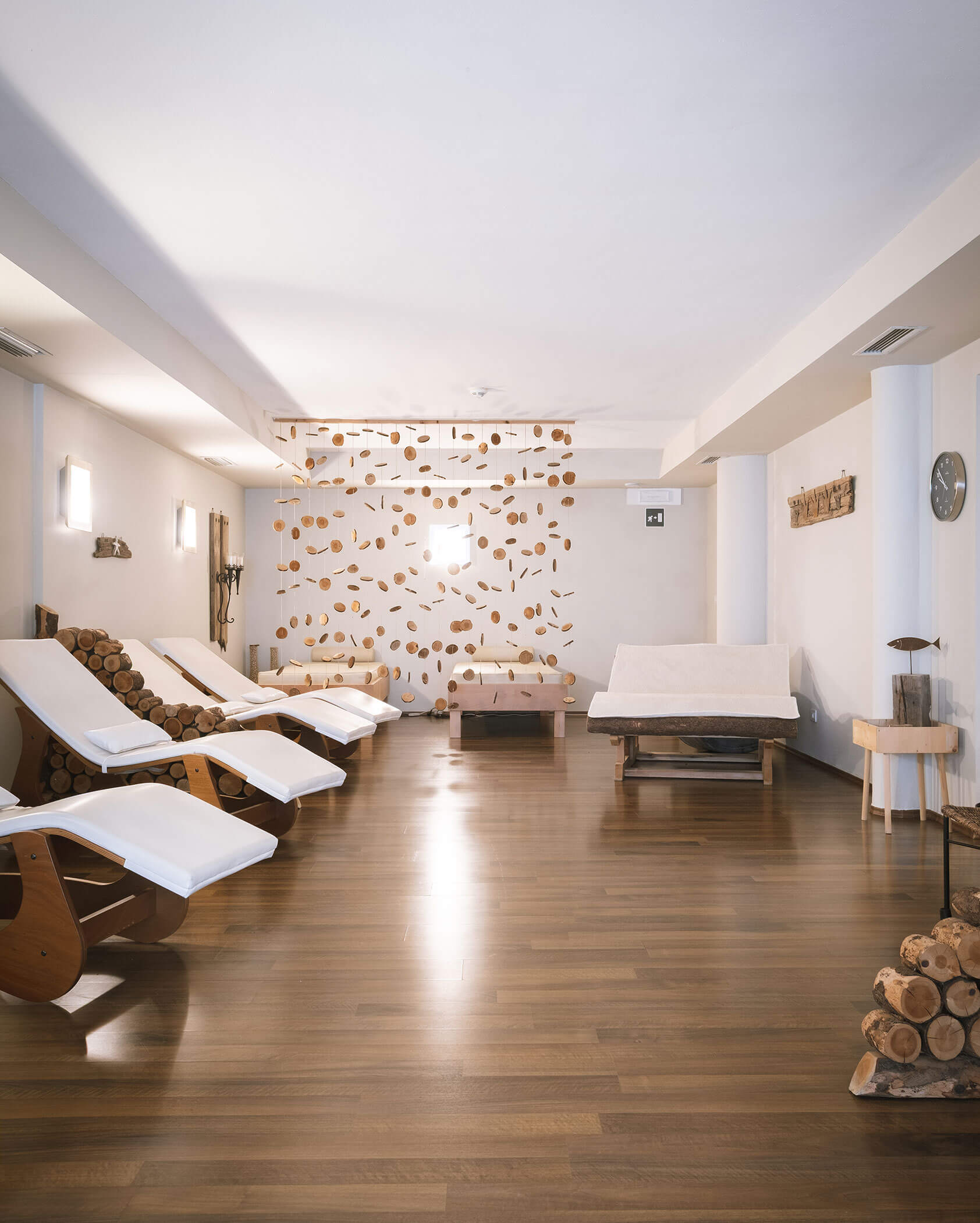 Moderna sala benessere con pavimenti in legno, sedie a sdraio bianche e tronchi impilati su una parete chiara e decorativa. - Hotel Innerhofer