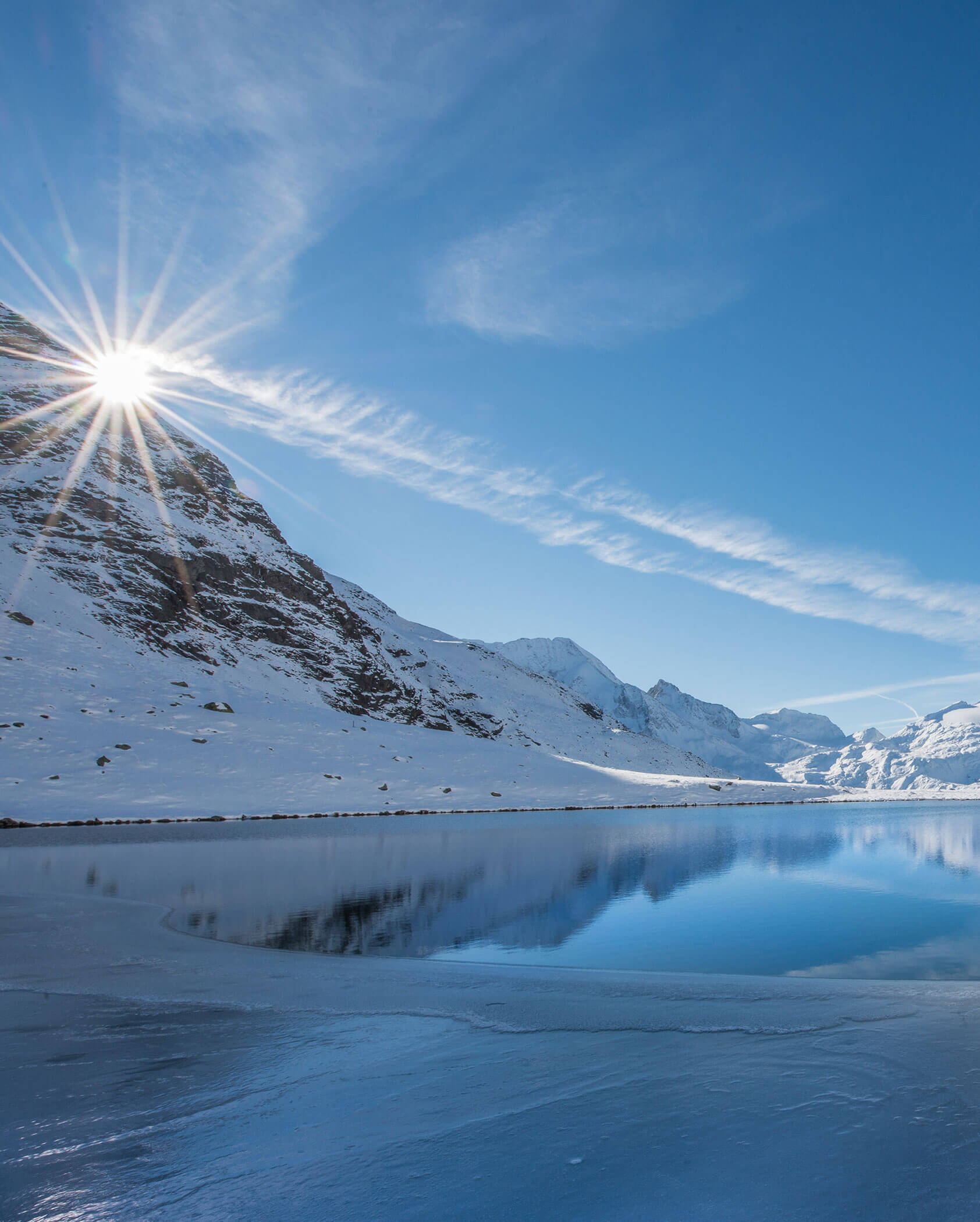 Il sole splende sulle montagne innevate, riflettendosi in un lago calmo e parzialmente ghiacciato sotto un cielo azzurro e limpido. - Hotel Innerhofer