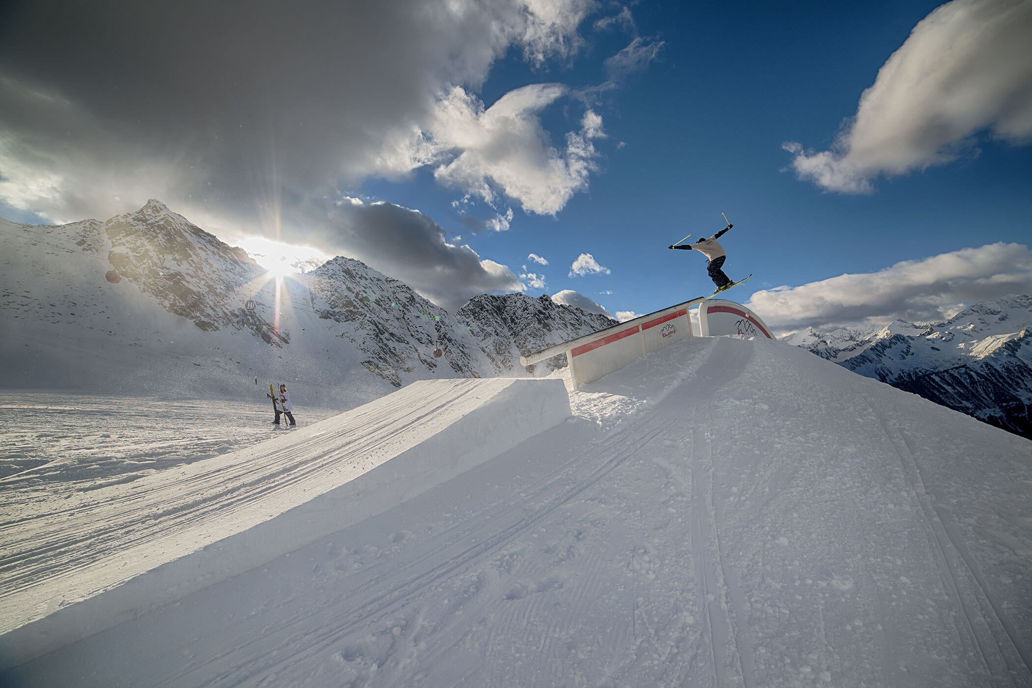 A snowboarder performs a trick on a rail in a snowy mountain terrain under a partly cloudy sky. - Hotel Innerhofer