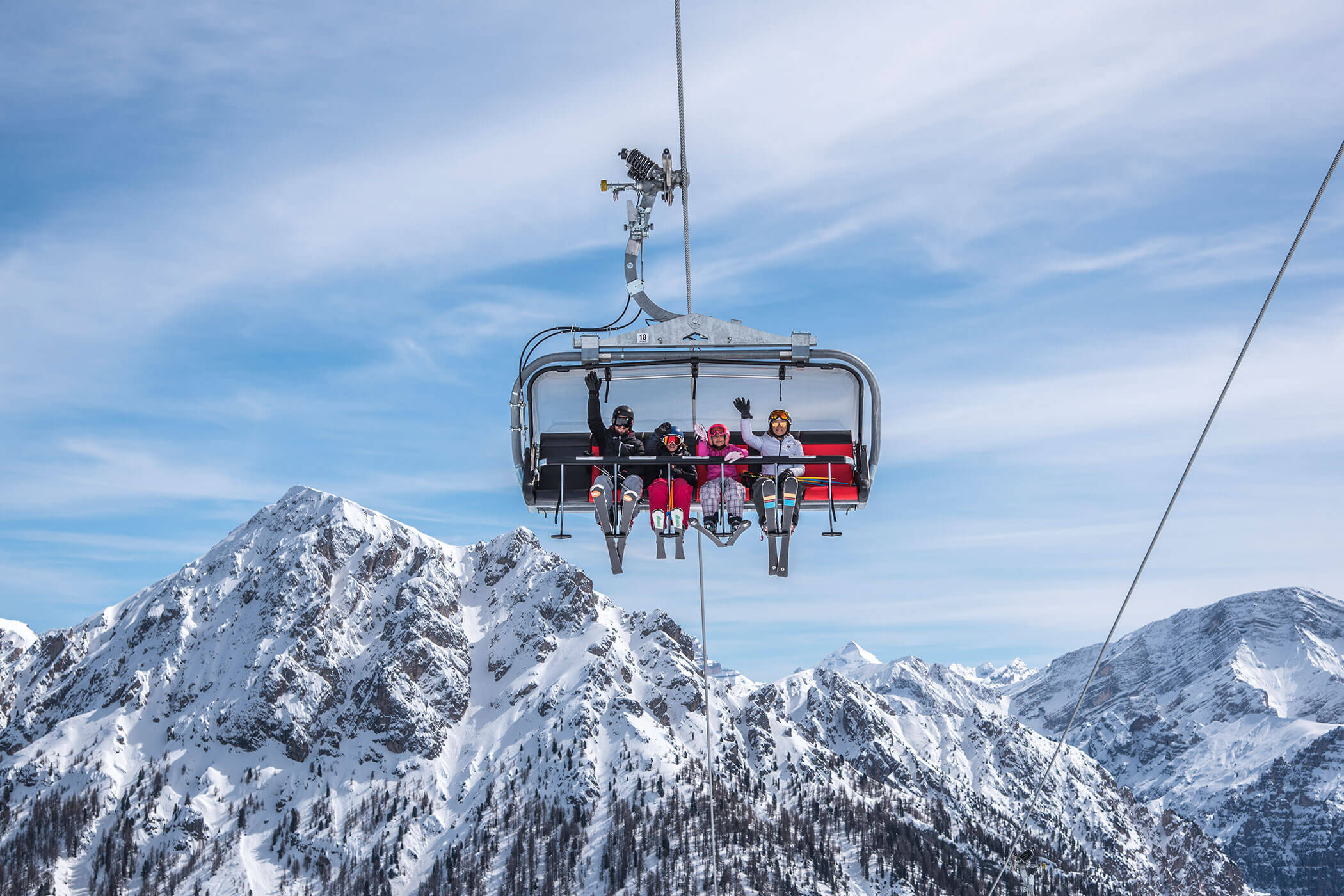 Four people riding a ski lift above snowy mountains under a blue sky with wispy clouds. - Hotel Innerhofer