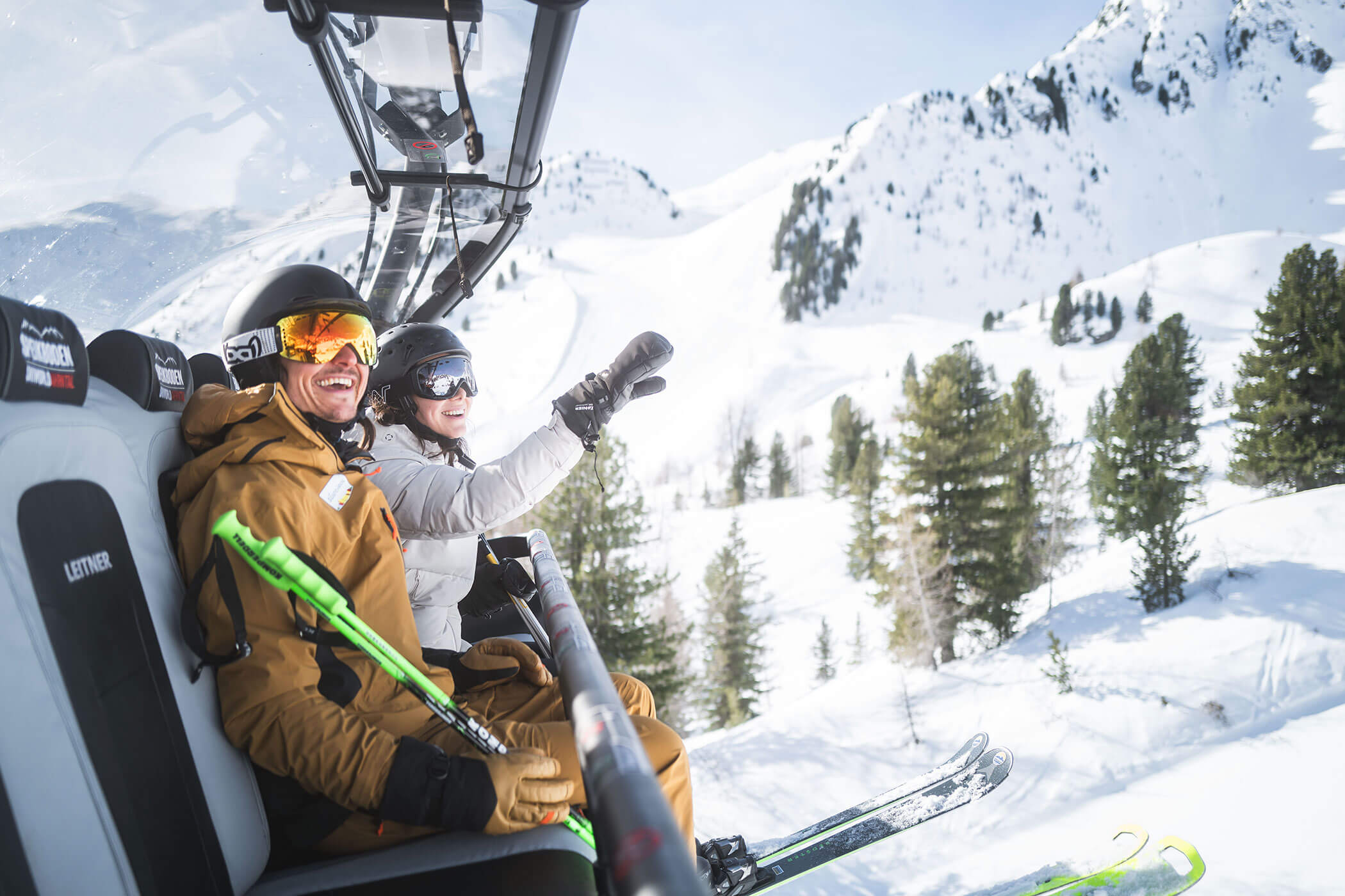 Two skiers on a ski lift, smiling and waving, with snowy mountains and pine trees in the background. - Hotel Innerhofer