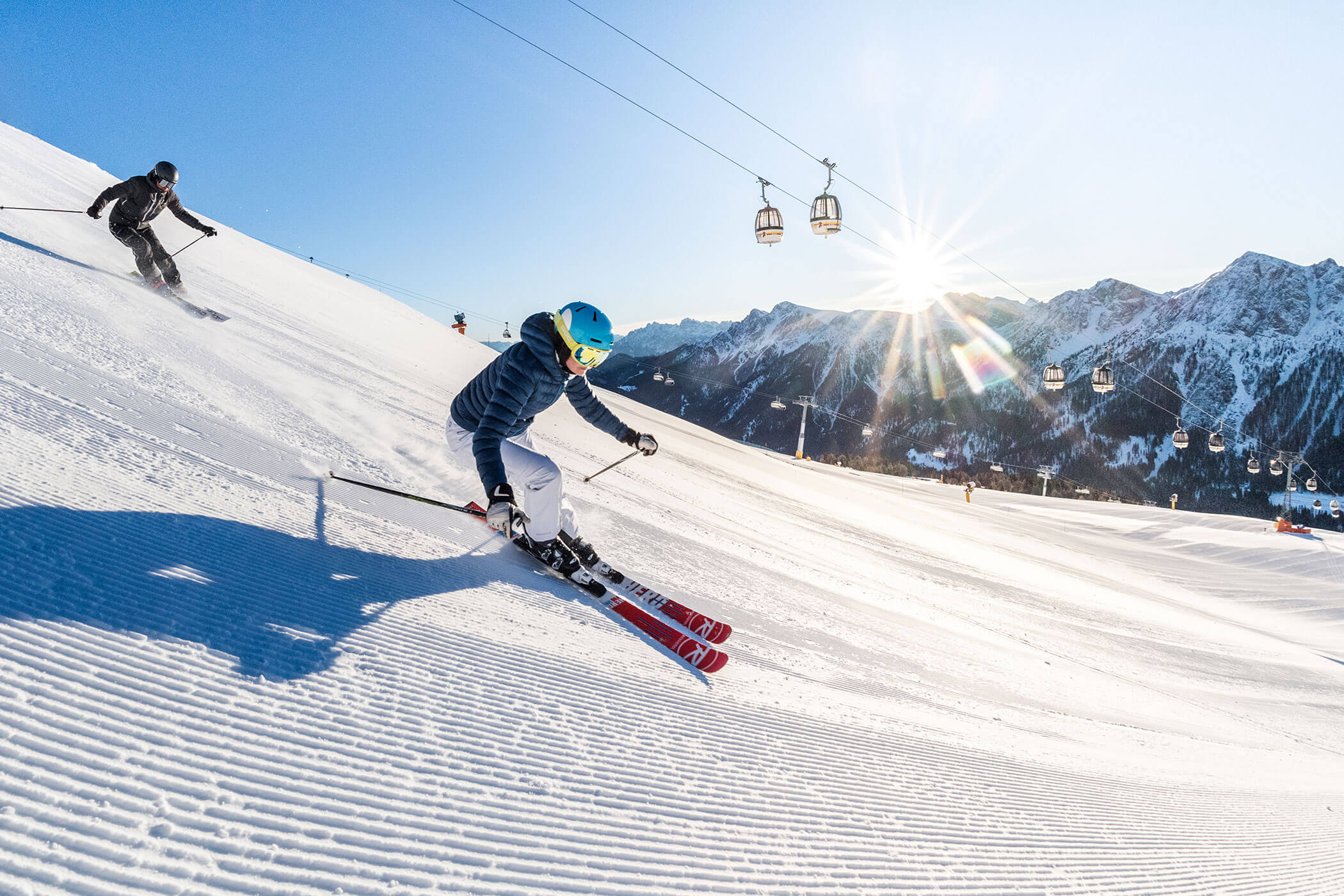 Two skiers descend a sunny, snowy slope with ski lifts and mountains in the background. - Hotel Innerhofer