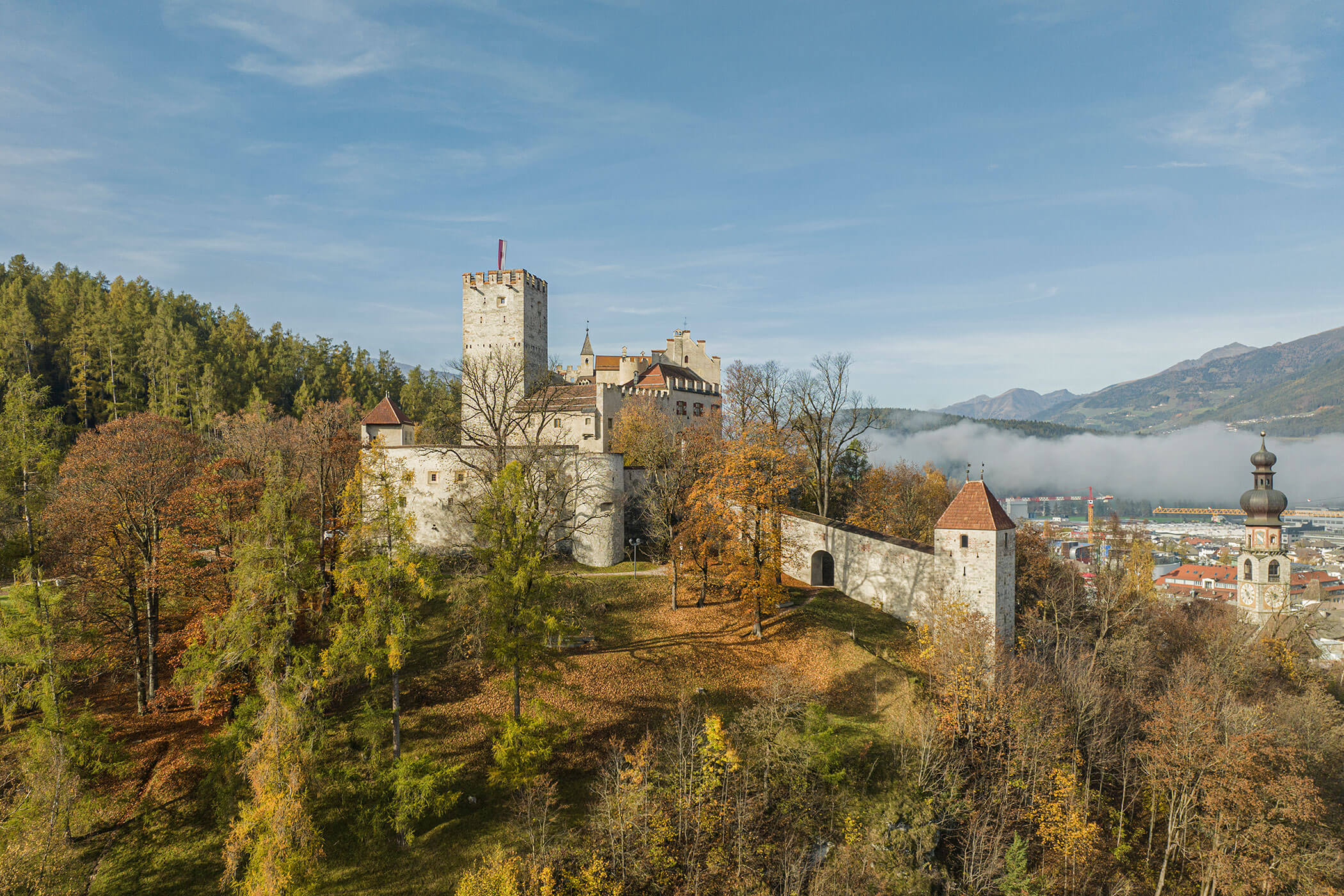 Castello medievale in pietra su una collina, circondato da alberi autunnali, con montagne e città sullo sfondo. - Hotel Innerhofer