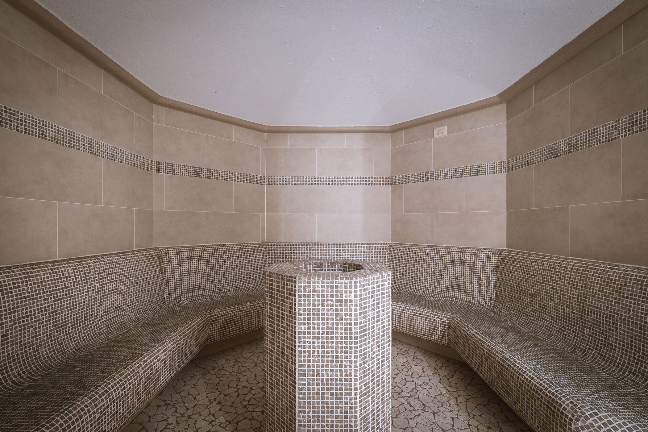 Bagno turco rivestito di piastrelle con panche curve e piedistallo rotondo al centro, pareti e pavimento beige. - Hotel Innerhofer