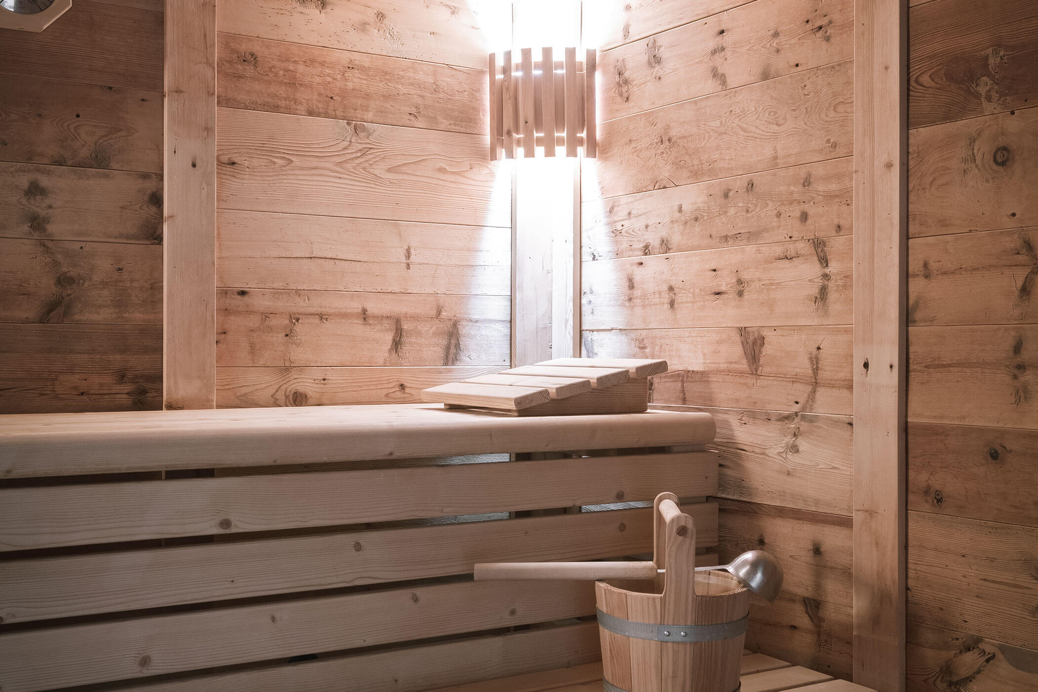 Interno della sauna in legno con panca, secchio e mestolo in legno e illuminazione soffusa sulla parete. - Hotel Innerhofer