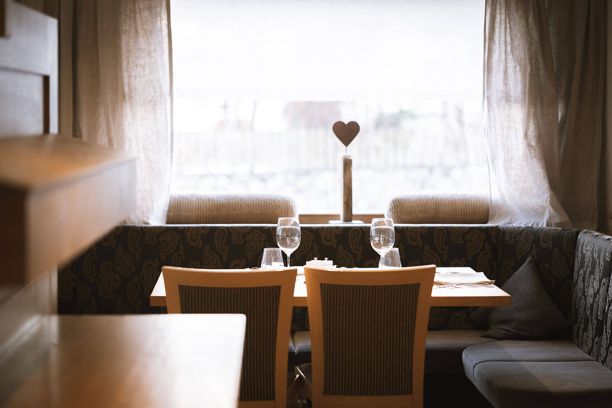 Gemütlicher Restaurantstand mit zwei Stühlen, gedecktem Tisch, Weingläsern und einer Herzdekoration am Fenster. - Hotel Innerhofer