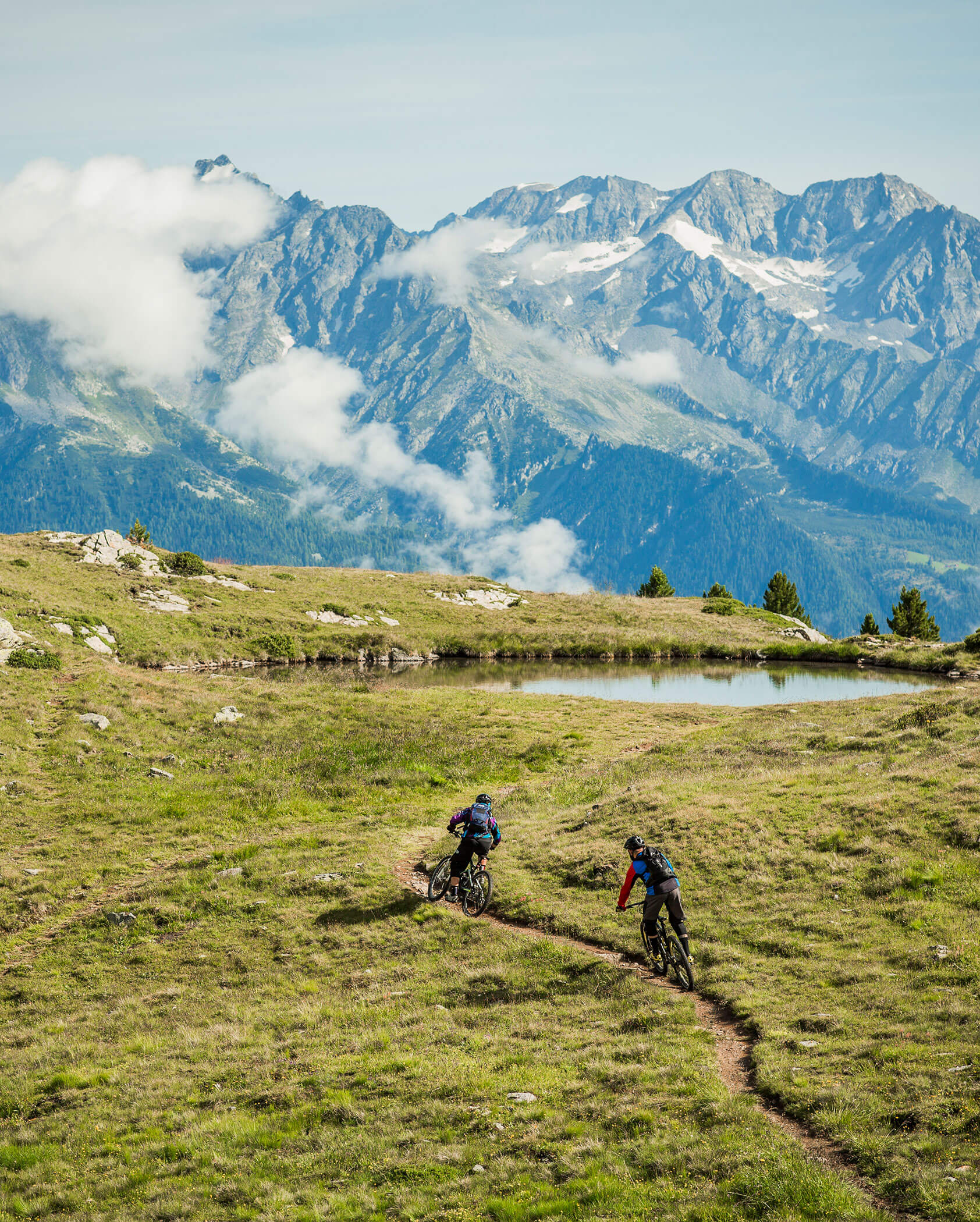 Two cyclists ride on a mountain trail with a scenic backdrop of tall, snow-capped peaks and grassy fields. - Hotel Innerhofer