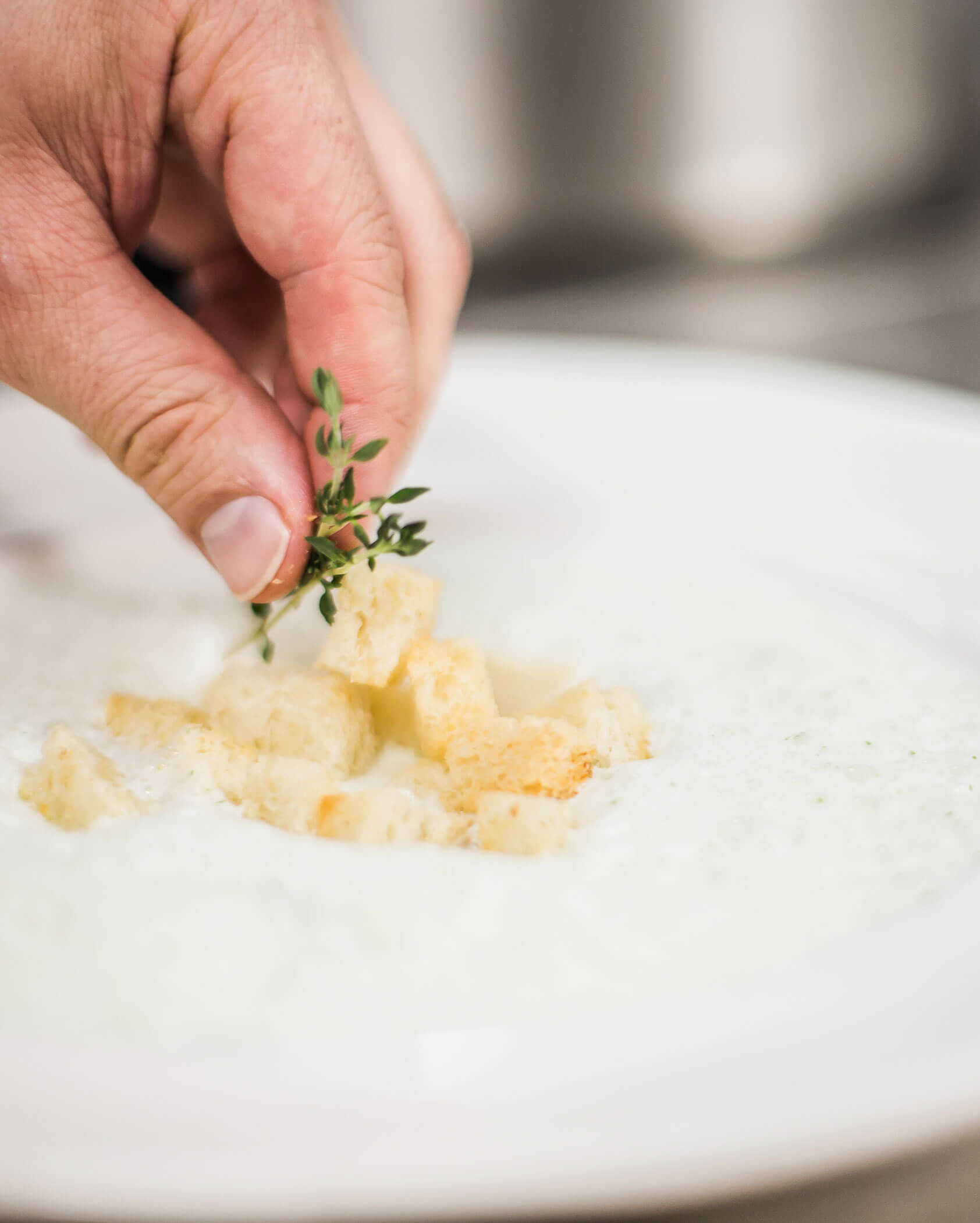 Hand garnishing creamy soup with croutons and a sprig of fresh thyme on a white plate. - Hotel Innerhofer