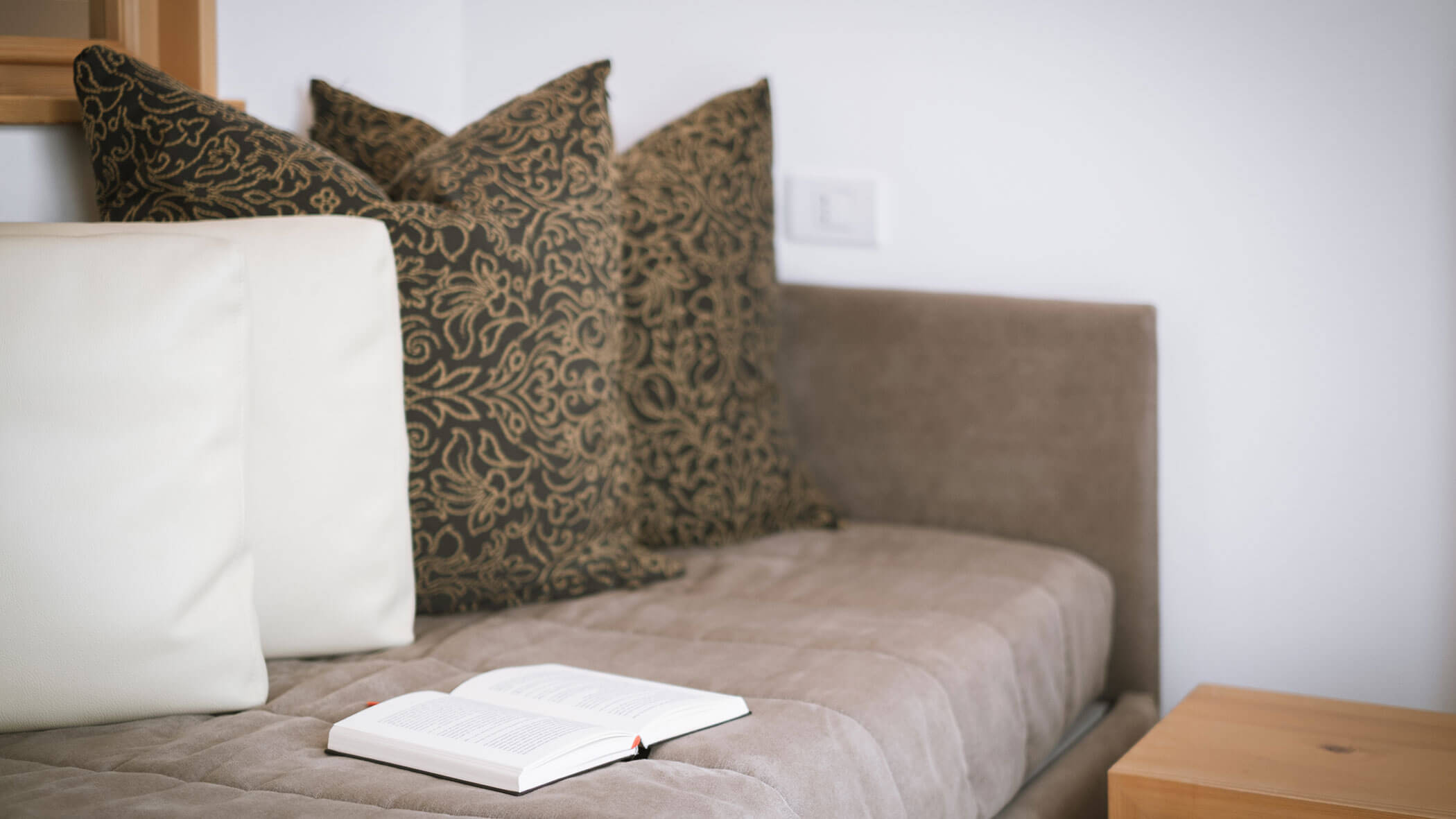 A cozy sofa with patterned pillows and an open book resting on the seat in a softly lit room. - Hotel Innerhofer
