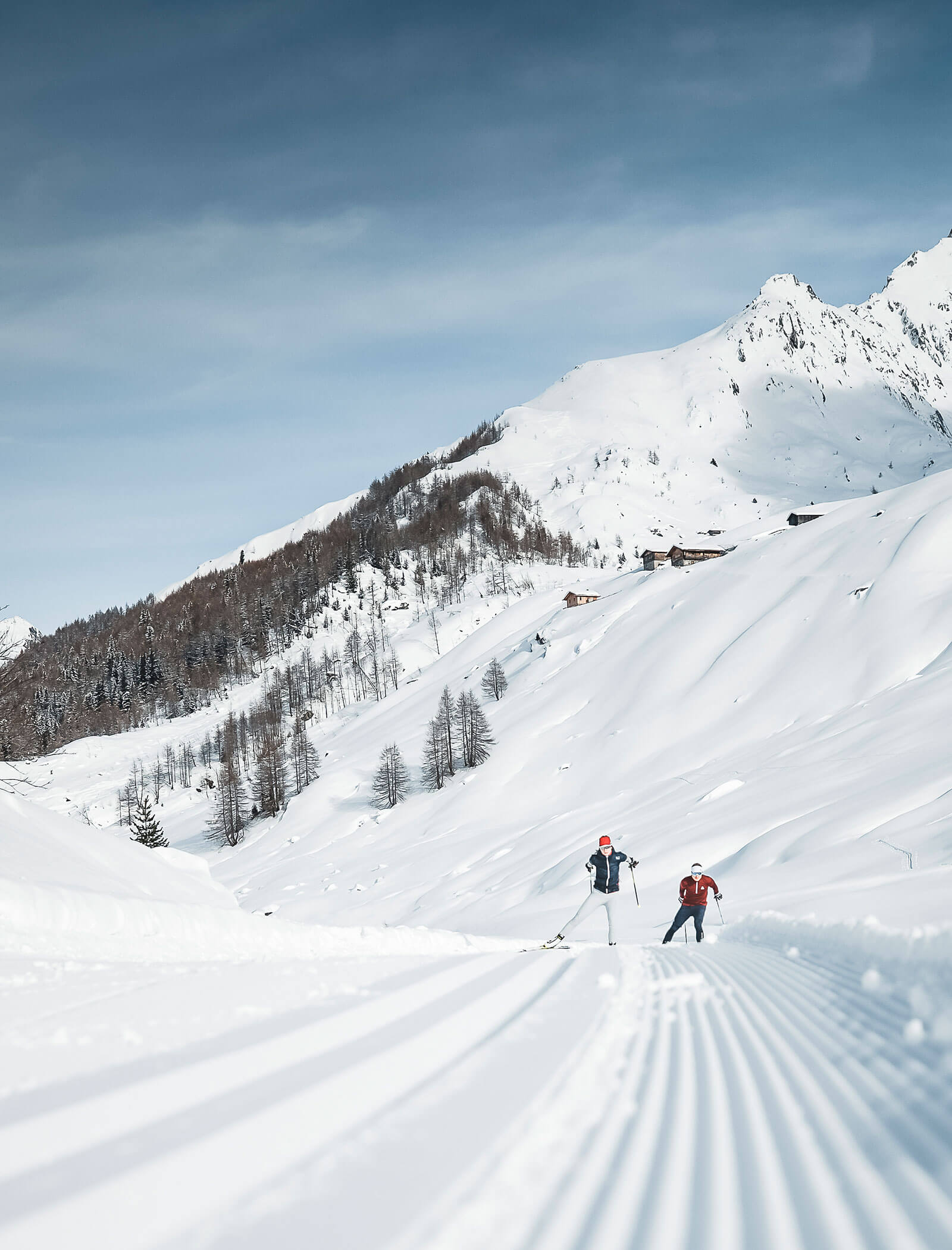 Two people skiing on a groomed snowy trail in a scenic mountain landscape under a blue sky. - Hotel Innerhofer