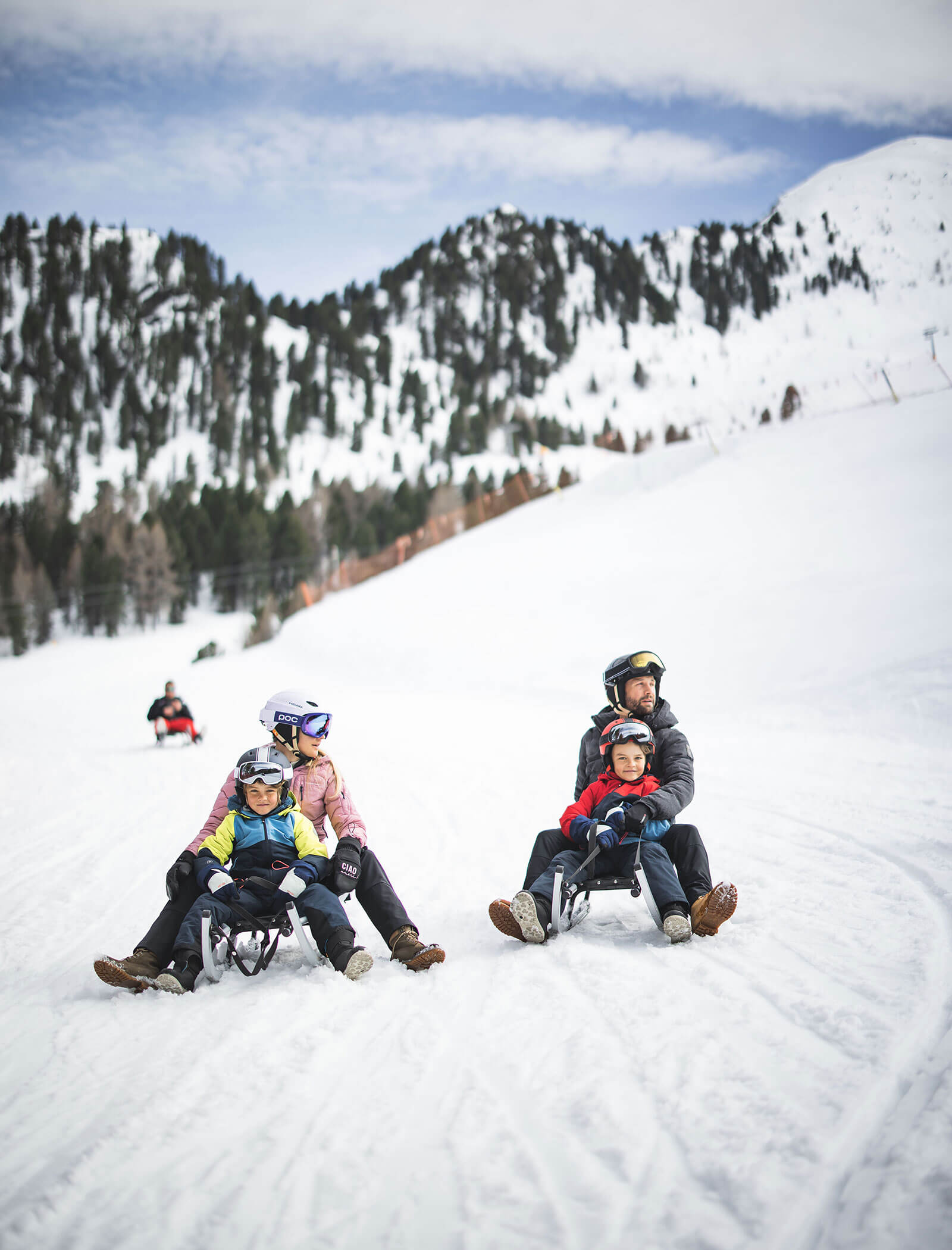 Two families sledding down a snowy mountain slope, surrounded by snow-covered trees and mountains. - Hotel Innerhofer