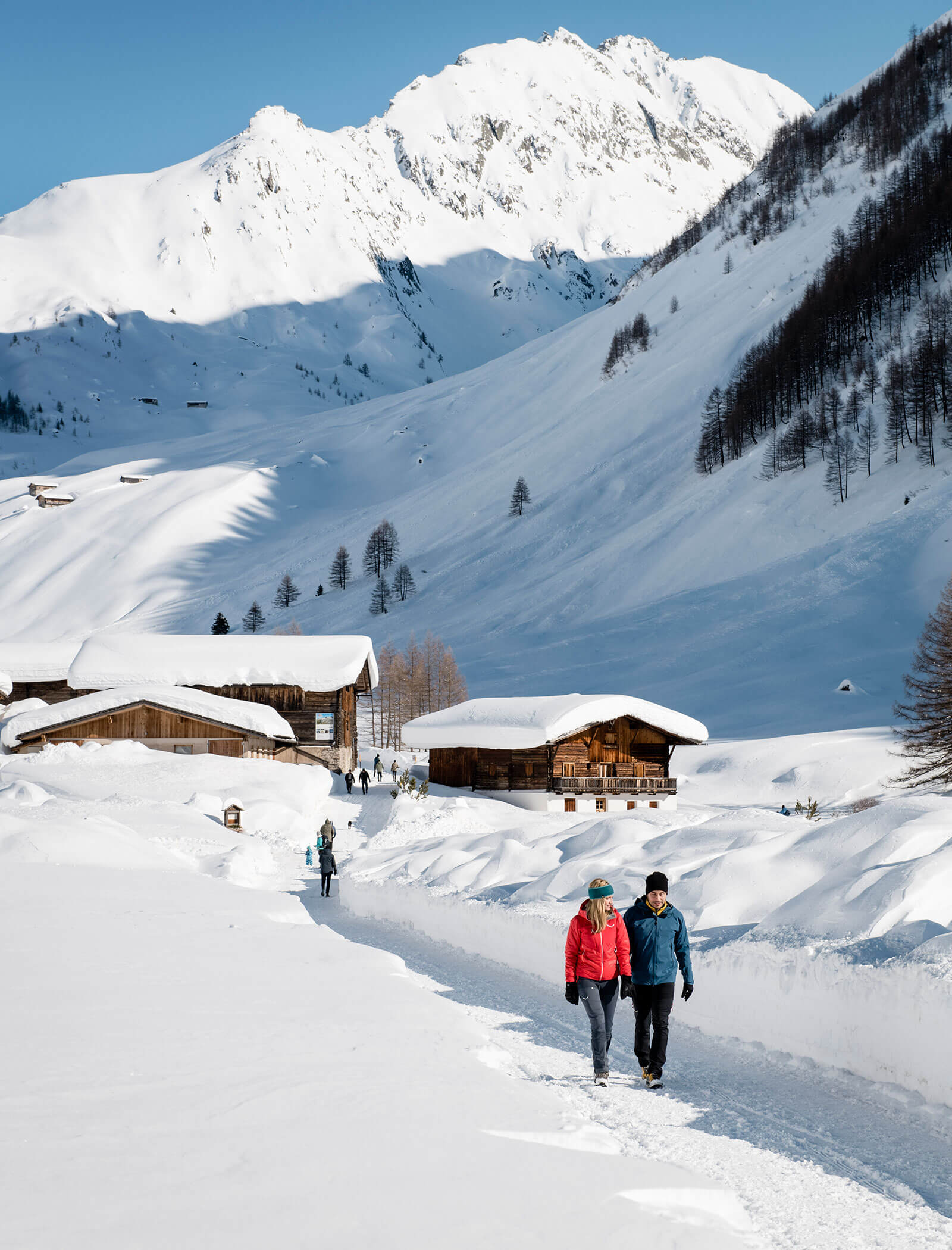 Two people walk on a snowy path near wooden cabins, with snow-covered mountains in the background. - Hotel Innerhofer