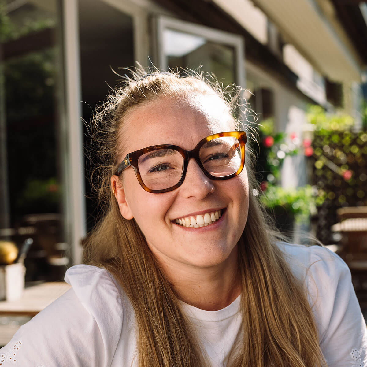 Lächelnde Frau mit langen blonden Haaren und großer Brille, die draußen im Sonnenlicht sitzt. - Hotel Innerhofer